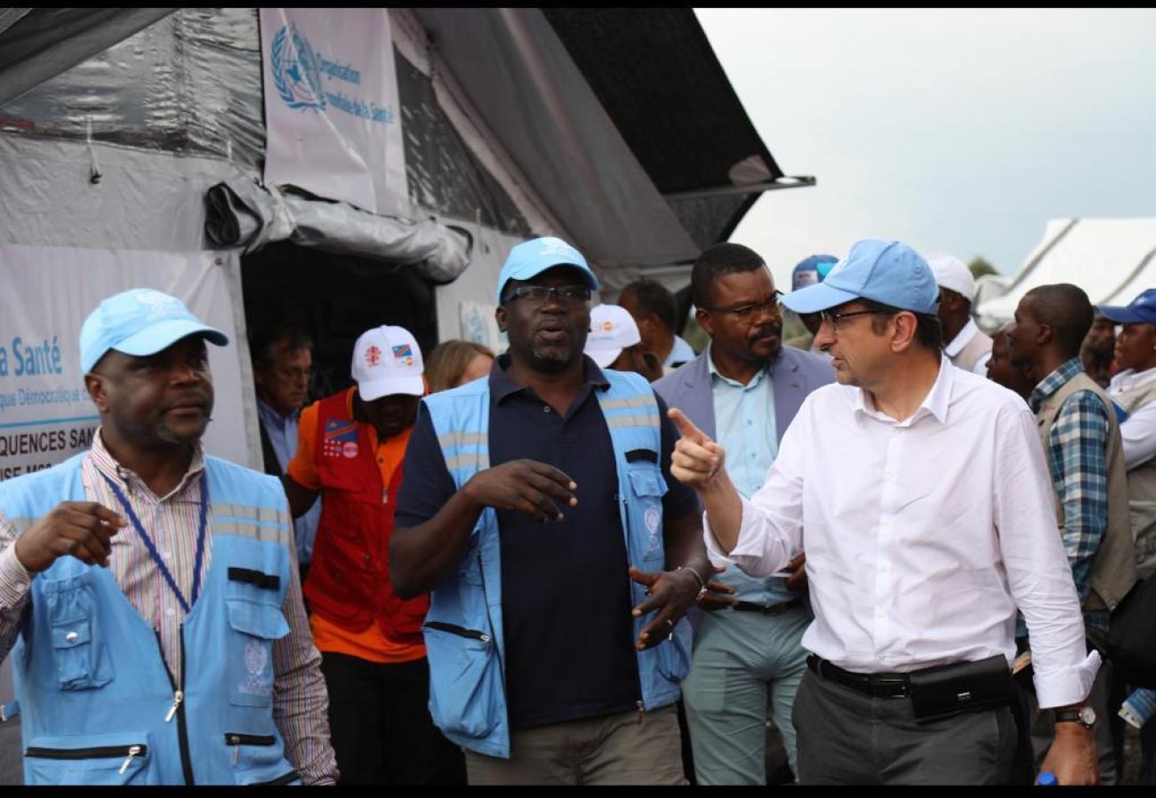 A man in a white shirt and blue cap talks to another man in a blue vest as other men stand in the background in the Democratic Republic of Congo