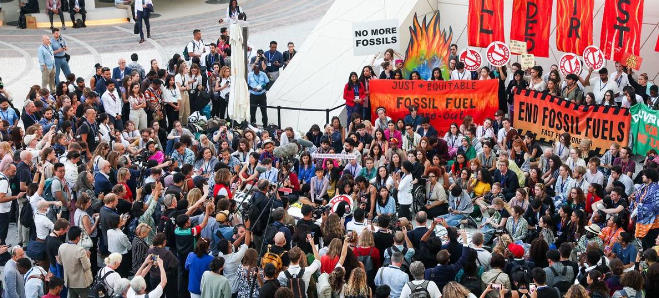 A crowd of protestors, carrying signs gather in front of a platform where people hold up a red poster demanding the end of fossil fuels