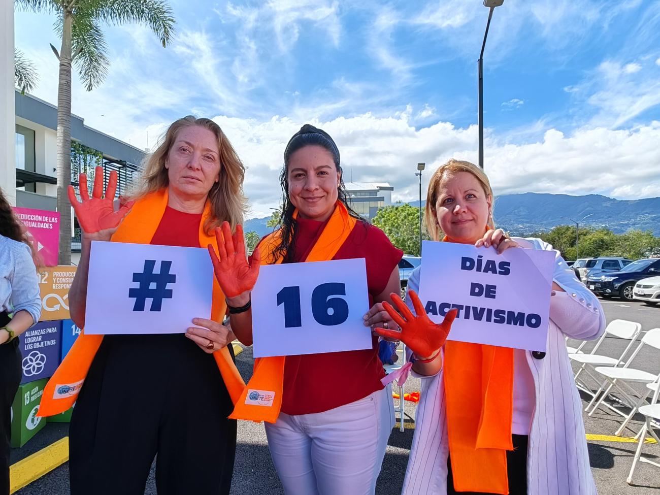 Three women, wear orange scarves and carry signs demanding an end to violence against women.