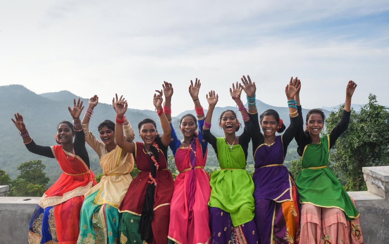 group of women wearing colourful saris smile to the camera with their hands in the air.