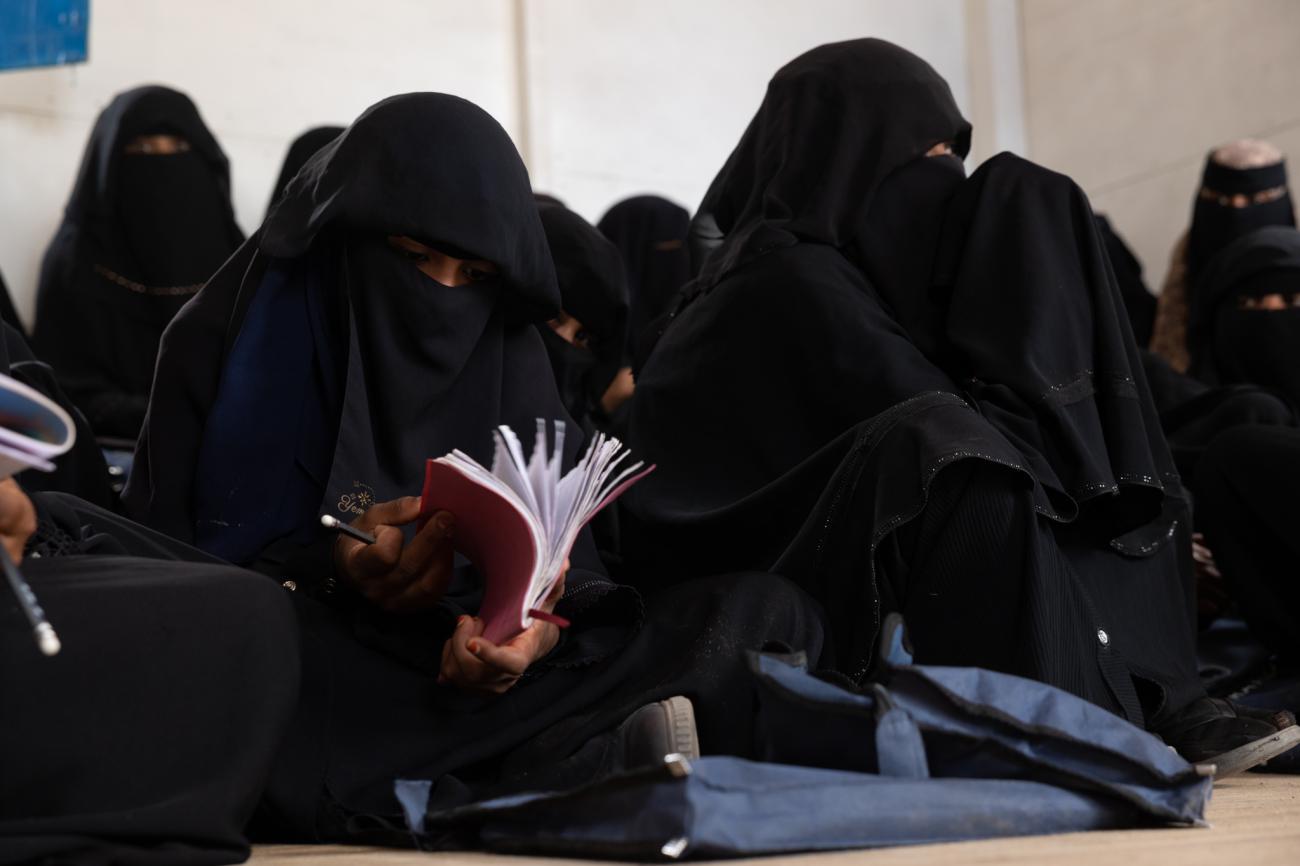 A row of women in full black clothing, sit on the floor with books laid out in front of them.