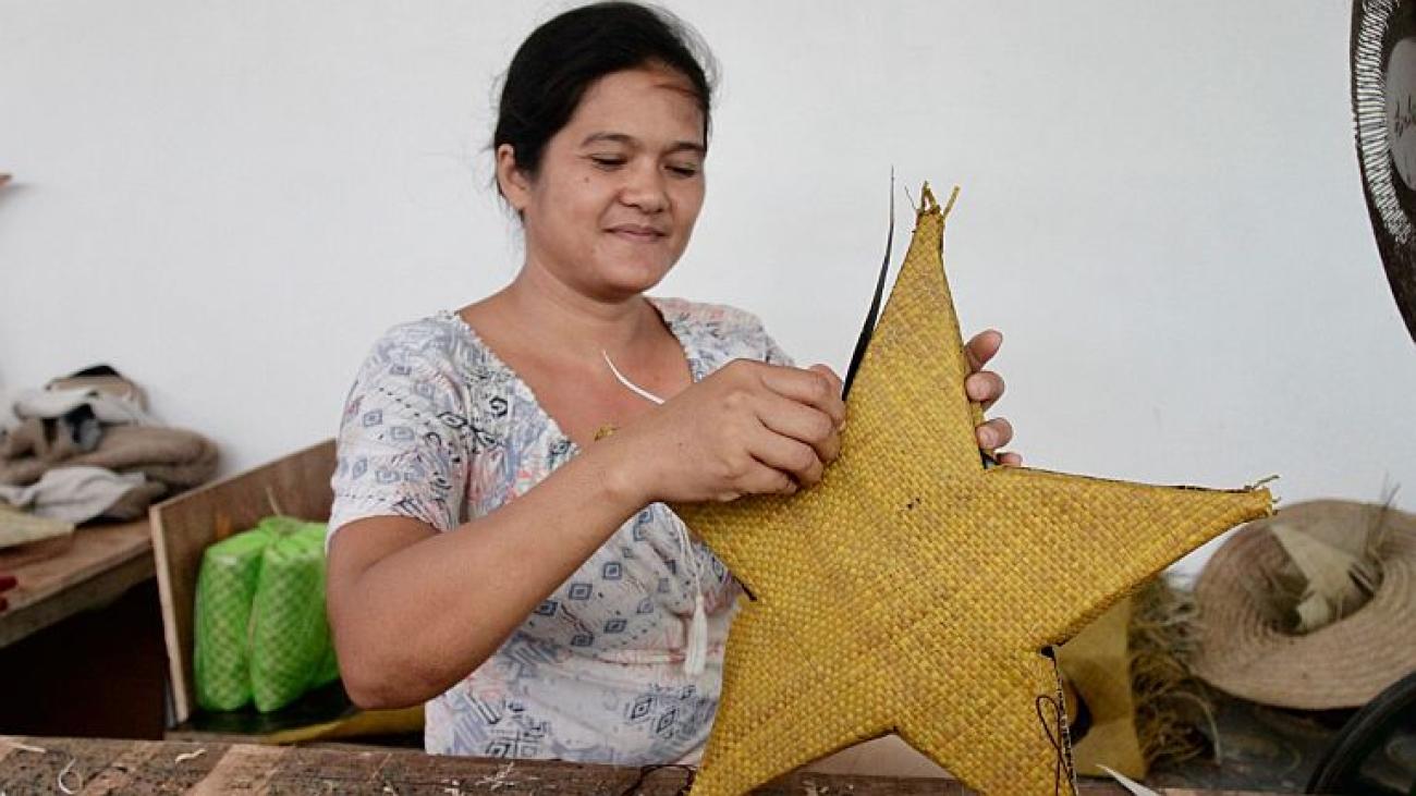 A woman in a white shirt weaves together a yellow star from reeds.