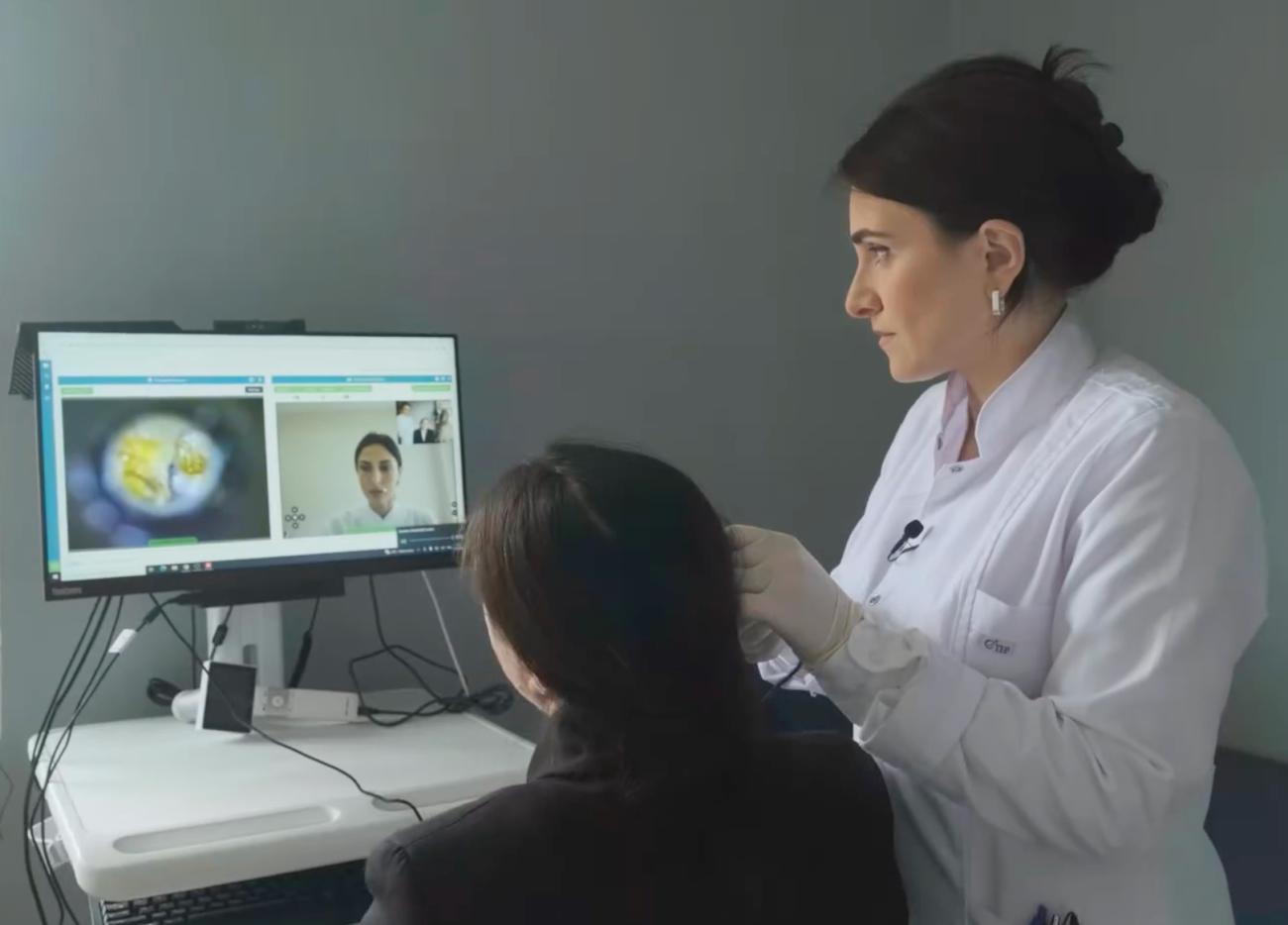 A doctor in a white coat diagnoses a woman in black clothing while another doctor is on a computer screen in front of them.