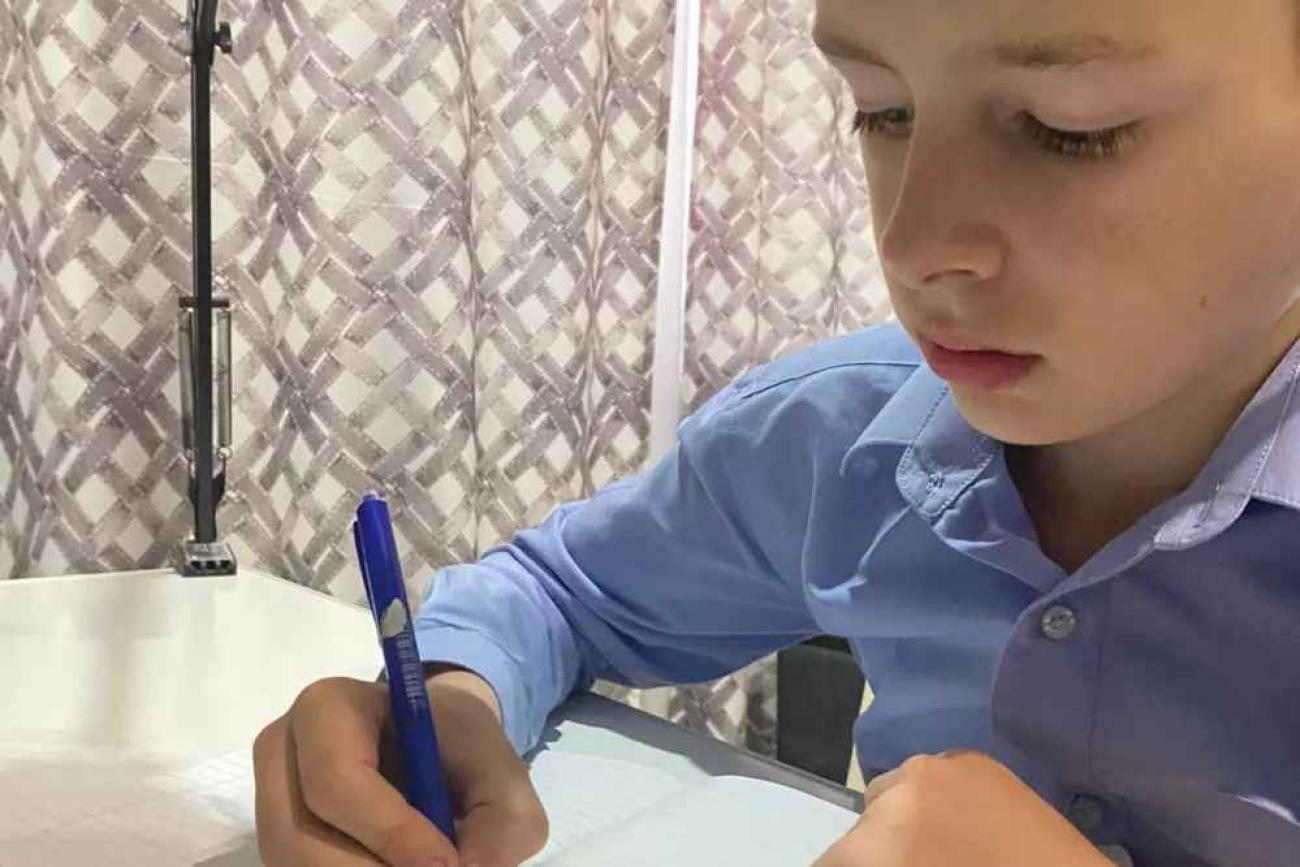 Мальчик в рубашке сидит за столом и пишет ручкой