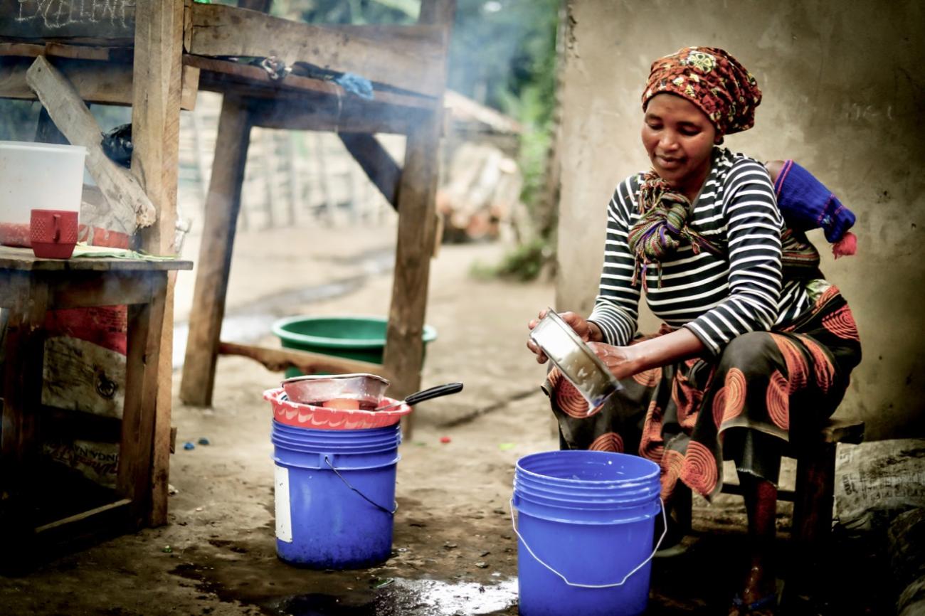 A woman sits wistfully and washes a metal pan/dish over buckets of water and soap.