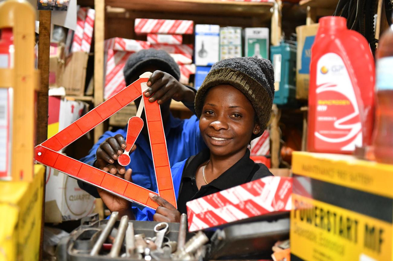 A woman in a headcap holds up a red sign inside a mechanic shed surrounded by tools