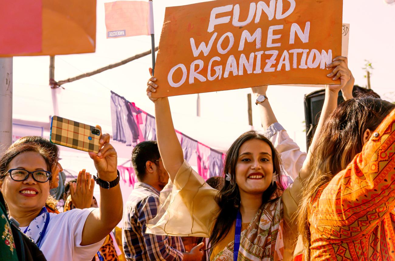 A group of women clad in orange taking part in a march, one of them holds up a sign saying "fund women's organizations"