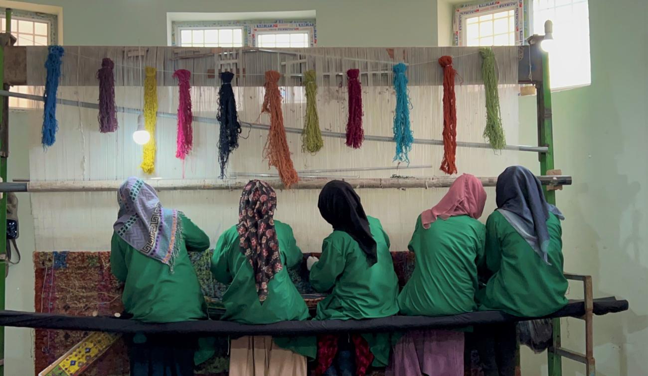 Women engaging in weaving, their heads are covered and their backs are turned to the camera