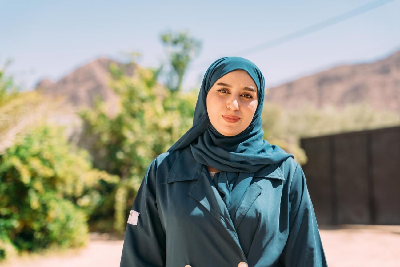 A woman in a teal dress and headscarf stands in the sun. Behind her are mountains and green vegetation