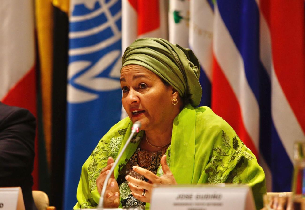A woman in a green dress and headscarf speaks into a microphone. She sits in front of a row of country flags.