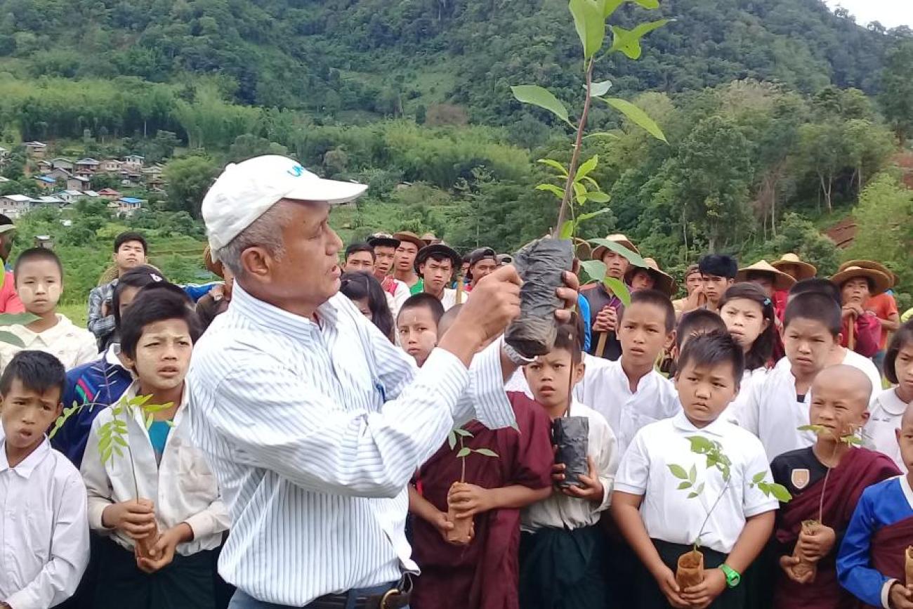 A man in a white shirt and white cap holds up a sapling in front of a group of school students.
