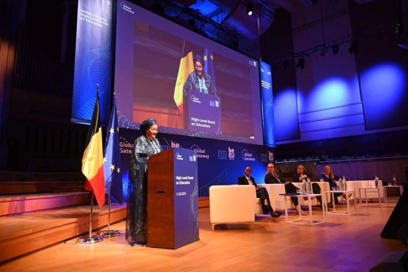 DSG Amina J. Mohammed stands behind a lectern and addresses an audience. She on stage with others who are seated at a panel. 