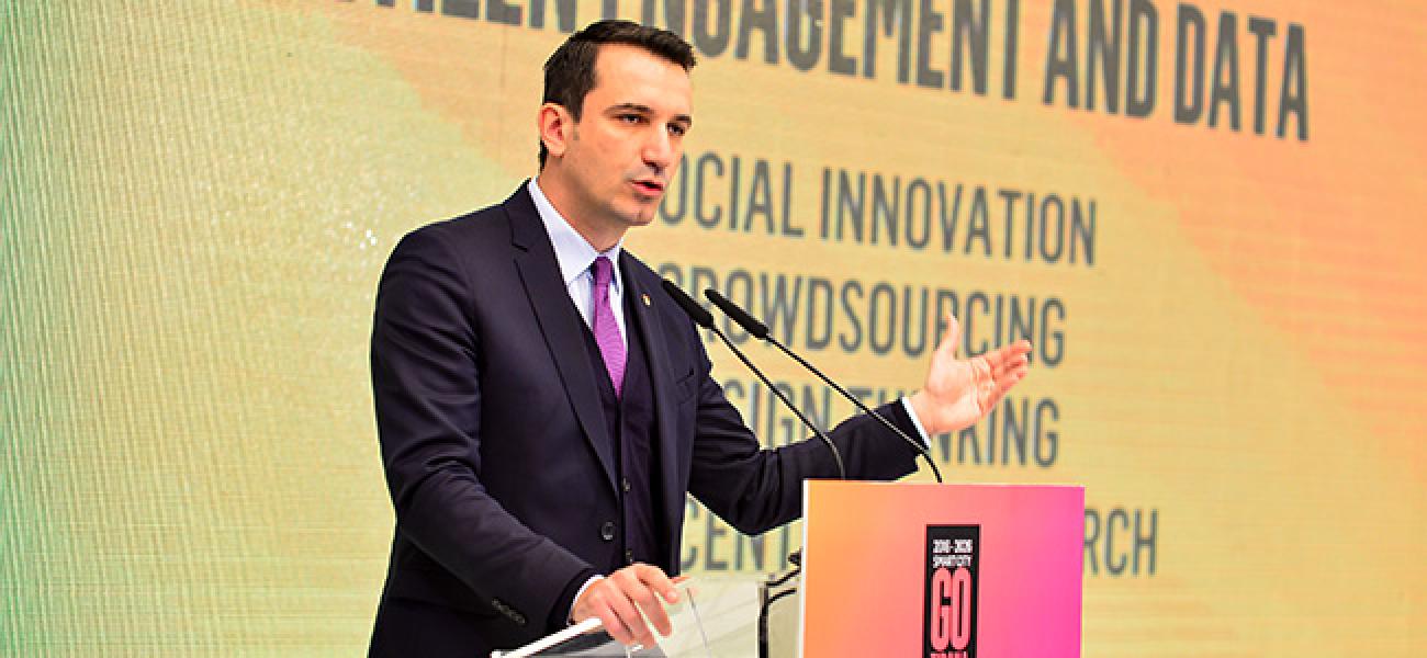 Un homme en costume cravate se tient debout derrière un pupitre et prononce un discours.