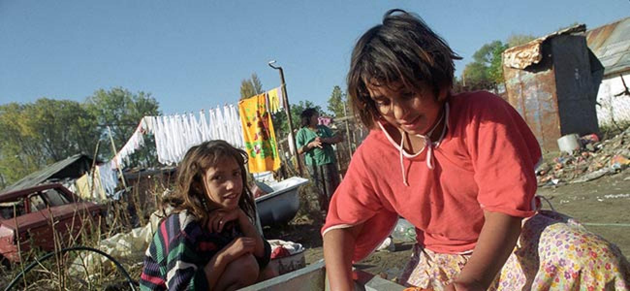 Deux adolescentes roms sont accroupies près d'une bassine, dans un espace découvert où l’on aperçoit, en arrière-plan, une femme et du linge étendu sur une corde.