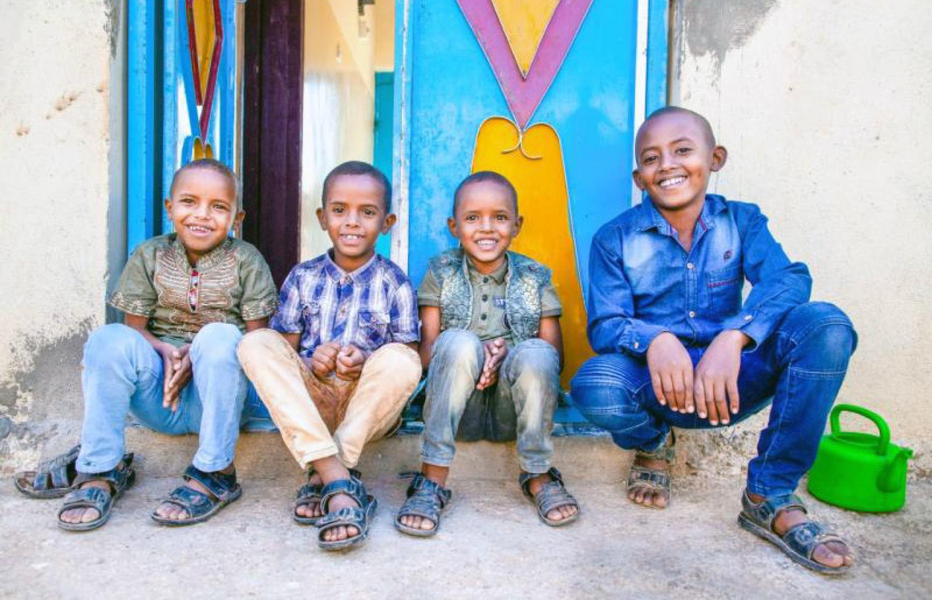 Four young boys sit outside on the step in front of an entrance. They all smile happily at the camera. 