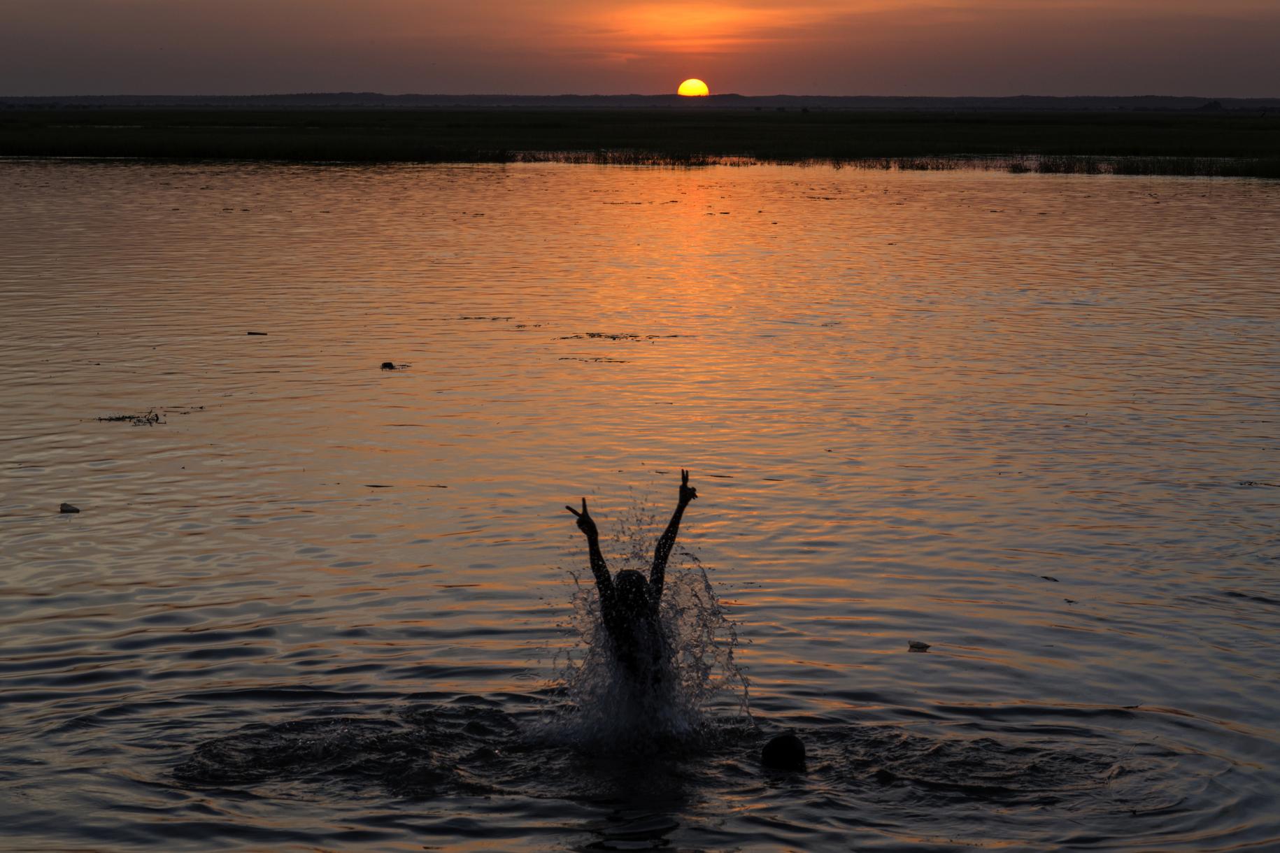 Una mujer salta del agua con los brazos en alto, mientras se pone el sol.