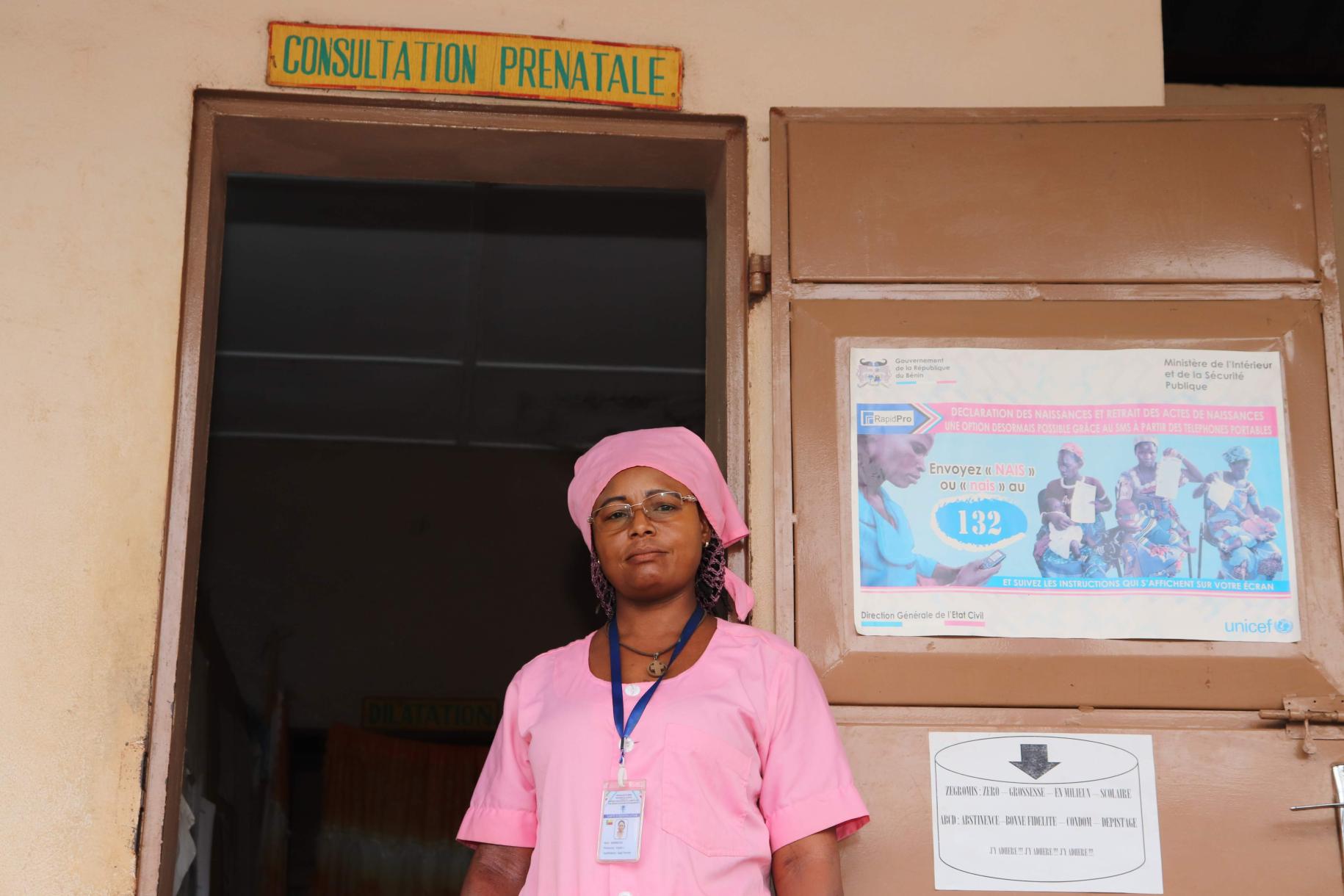 Una mujer con un uniforme rosa y una red que le cubre el cabello, está parada frente a una puerta de un centro de salud. Se aprecia un letrero sobre la puerta donde se indica que se realizan consultas prenatales.