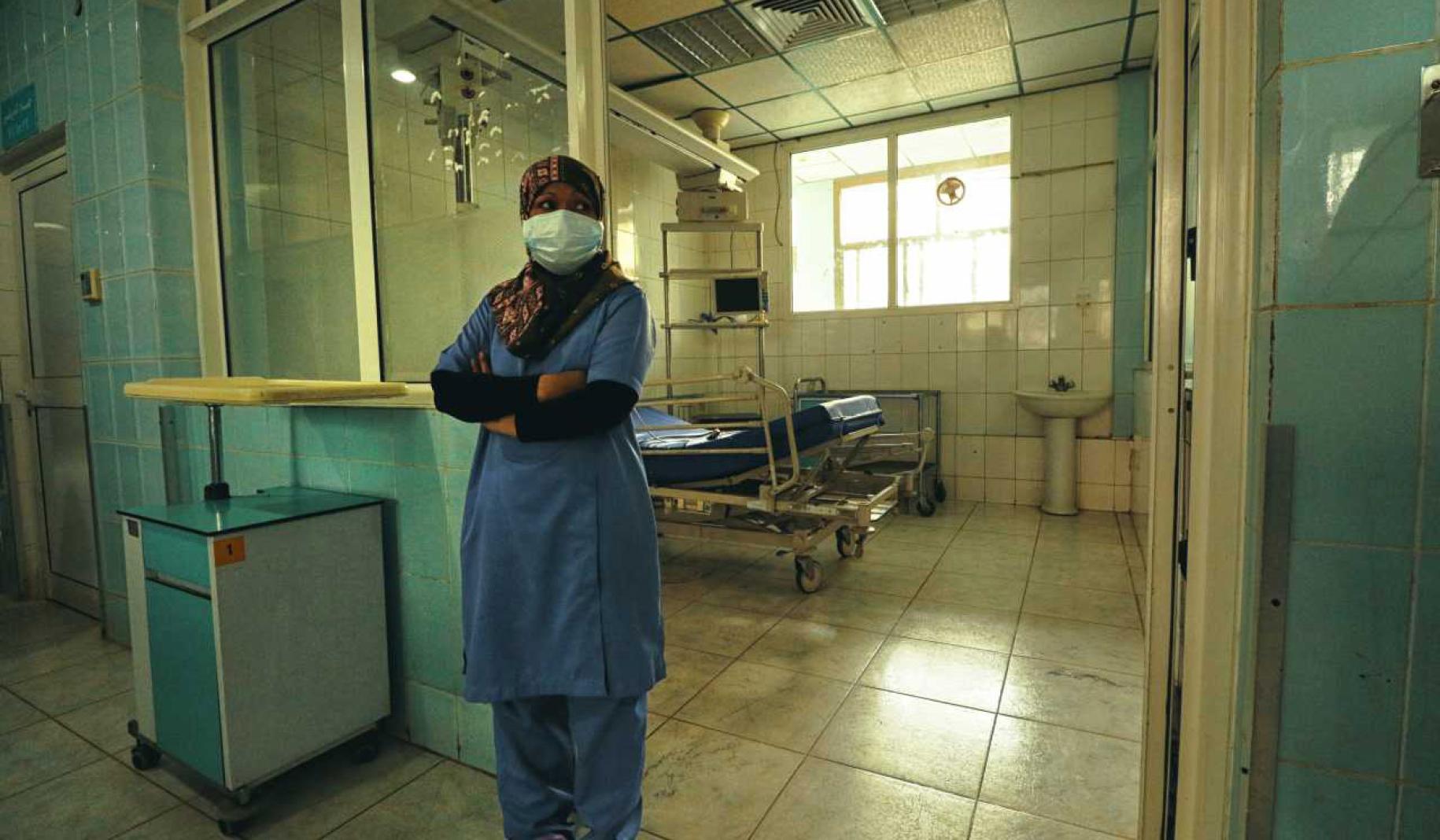Une infirmière se tient debout, les bras croisés, devant une chambre de l'hôpital Al Kuwait à Sana'a, au Yémen. Elle porte une blouse bleue et un masque de protection.