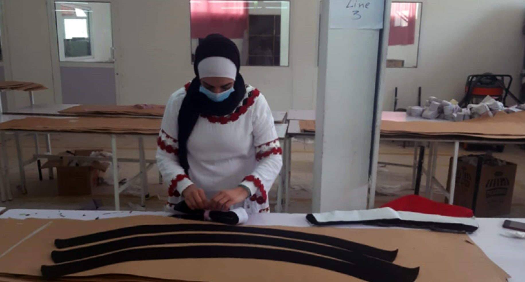 A woman wearing a protective face mask concentrates on a garment in a manufacturing space.