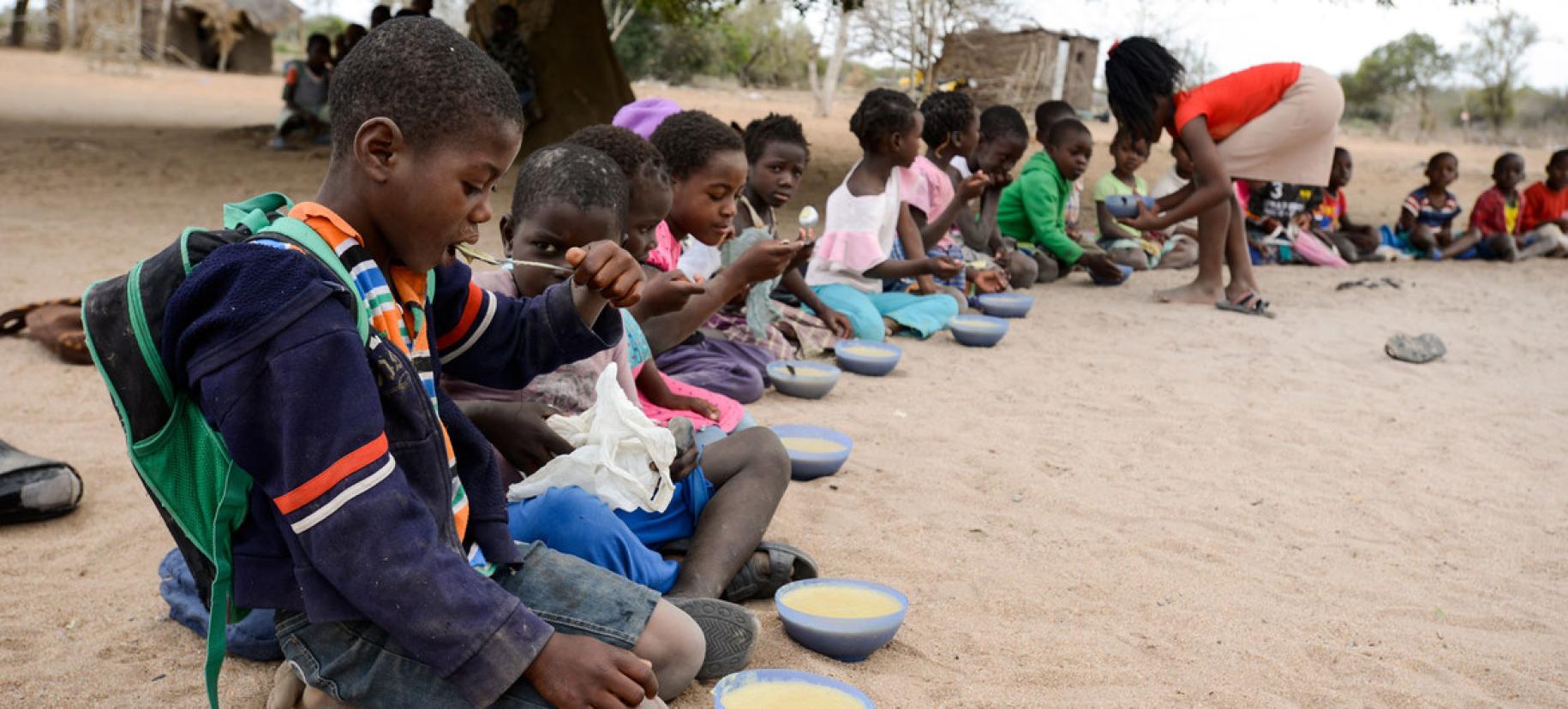 De jeunes enfants sont assis par terre, en demi-cercle, à l'extérieur et prennent un repas que leur sert une jeune fille dans des assiettes bleues.