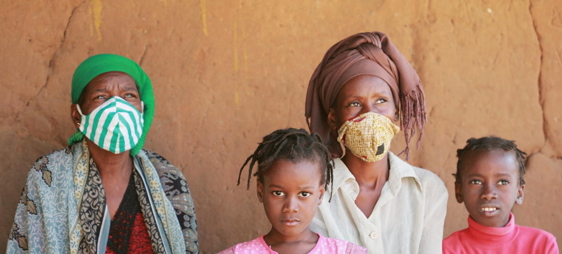 Deux femmes et deux enfants se tiennent face à la caméra. Les deux femmes portent un turban et un masque de protection respiratoire en tissu et regardent au loin. La fillette au centre de l'image regarde fixement l'appareil photo.