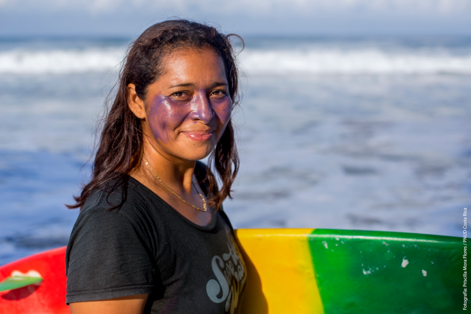 A female surfer smiles as she holds her surfboard by the shore of a beach.