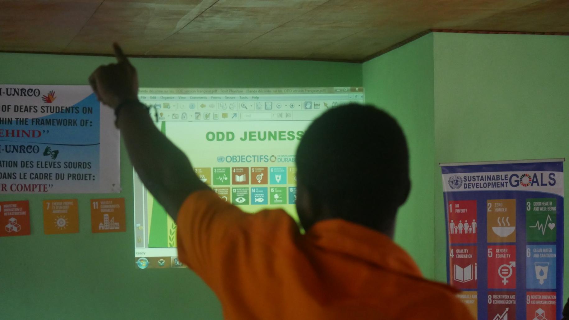 A young boy faces away from the camera is is raising a finger in front a projected screen showing the SDG icons.