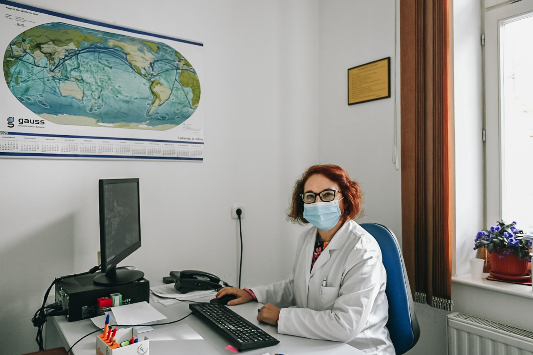 Dr. Snežana sitting at her desk with a poster of a world map just above her desk.
