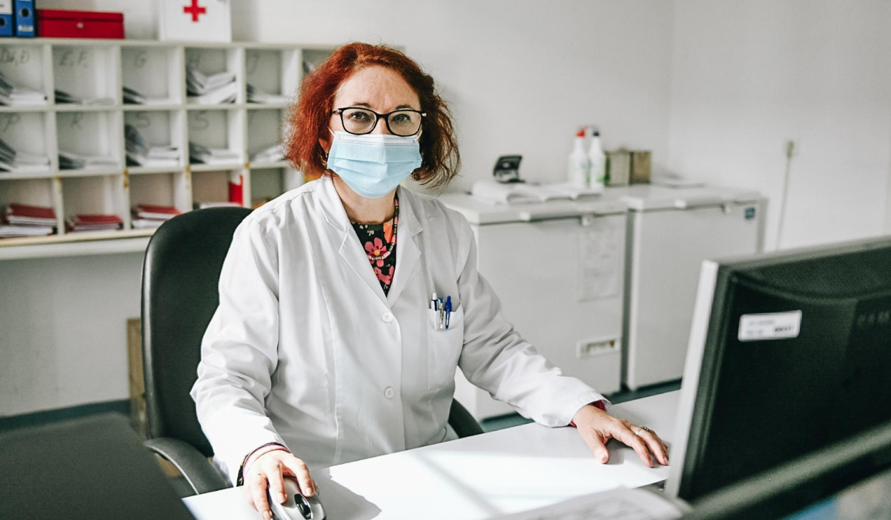 Snežana Bursać Aranđelović est assise à son bureau, un ordinateur devant elle, à l'Institut de santé publique du canton de Sarajevo. Elle porte une blouse blanche et un masque chirurgical bleu et elle regarde la caméra. On aperçoit derrière des dossiers et objets divers posés sur des étagères blanches.