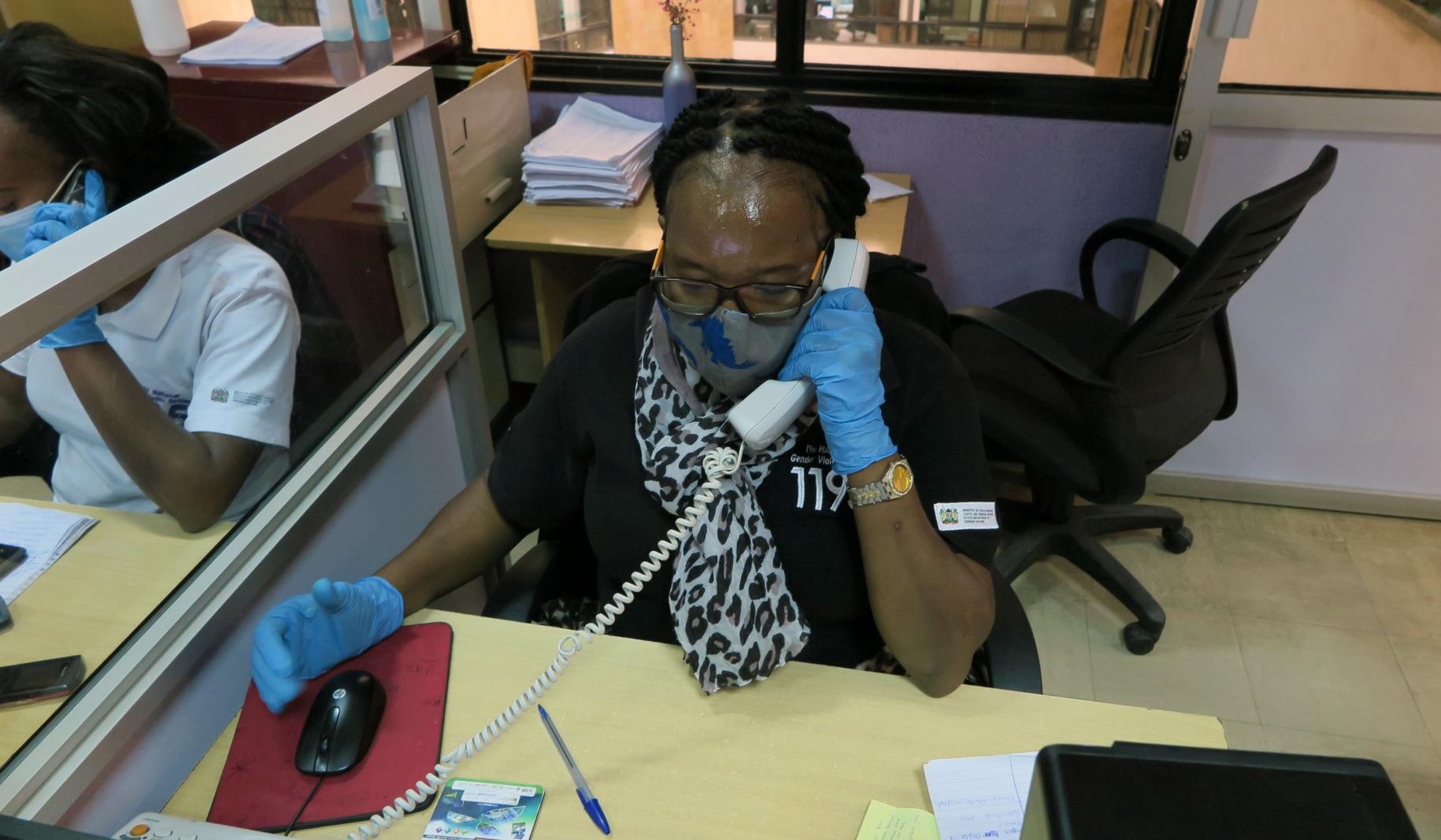 Una representante del centro de llamadas responde a una llamada realizada a la plataforma telefónica del servicio de atención a víctimas. Ella se sienta en un escritorio y usa una mascarilla y guantes protectores.