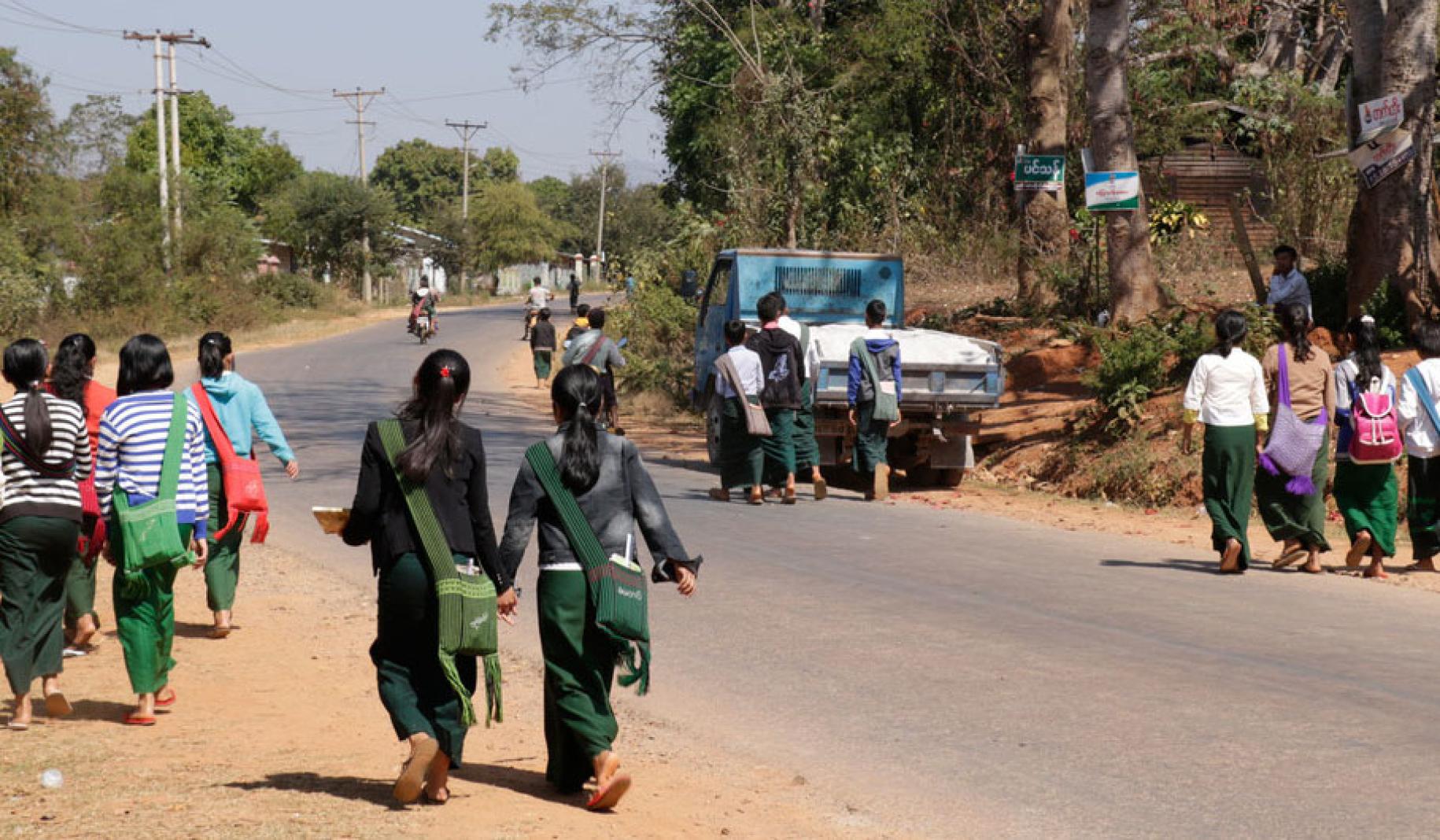 Alumnos caminando en grupo por el hombrillo de tierra de una carretera, en dirección a la escuela.