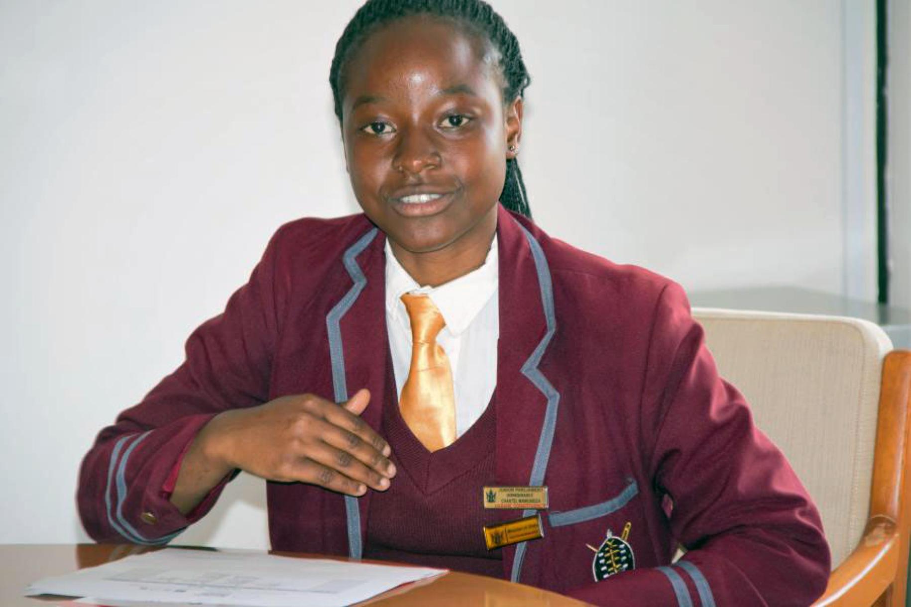 A young Zimbabwean school girl wearing her school uniform sits at a desk as she speaks.