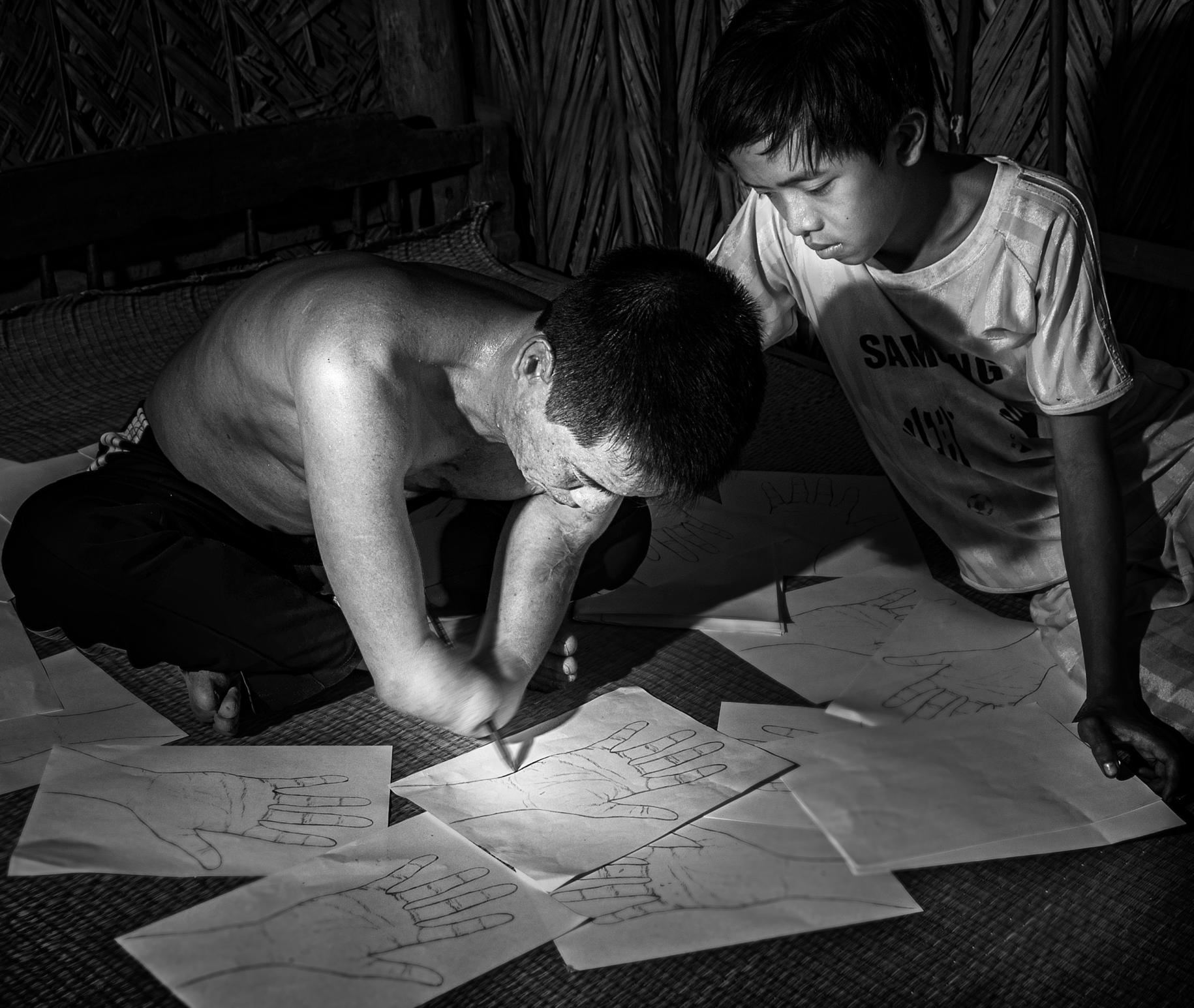 Black and white photo of a man and boy. The man has an amputated hand, sit together on the ground drawing and writing on sheets of paper.