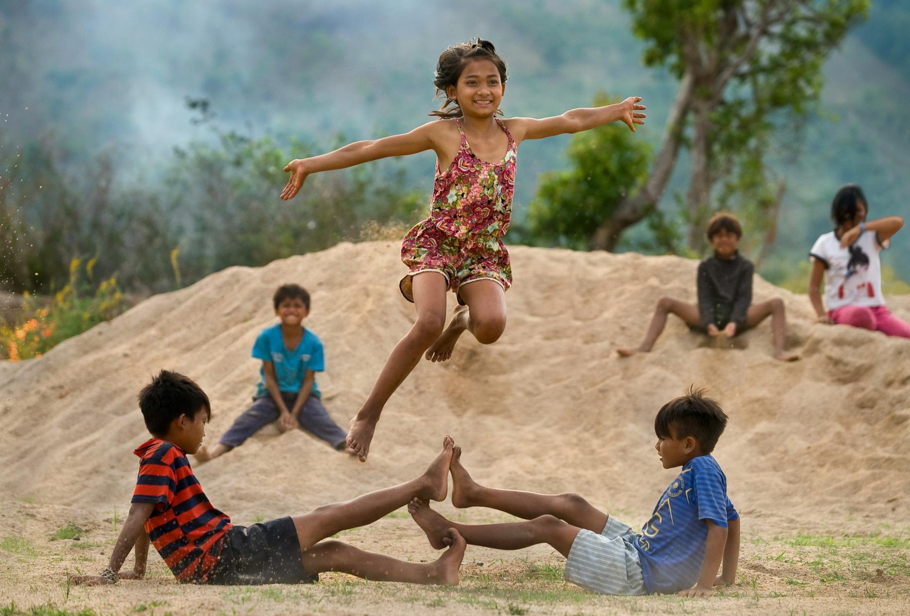 Children play outside. Two boys are on the ground touching their feet, while one girl is frozen in the air happily jumping above them.