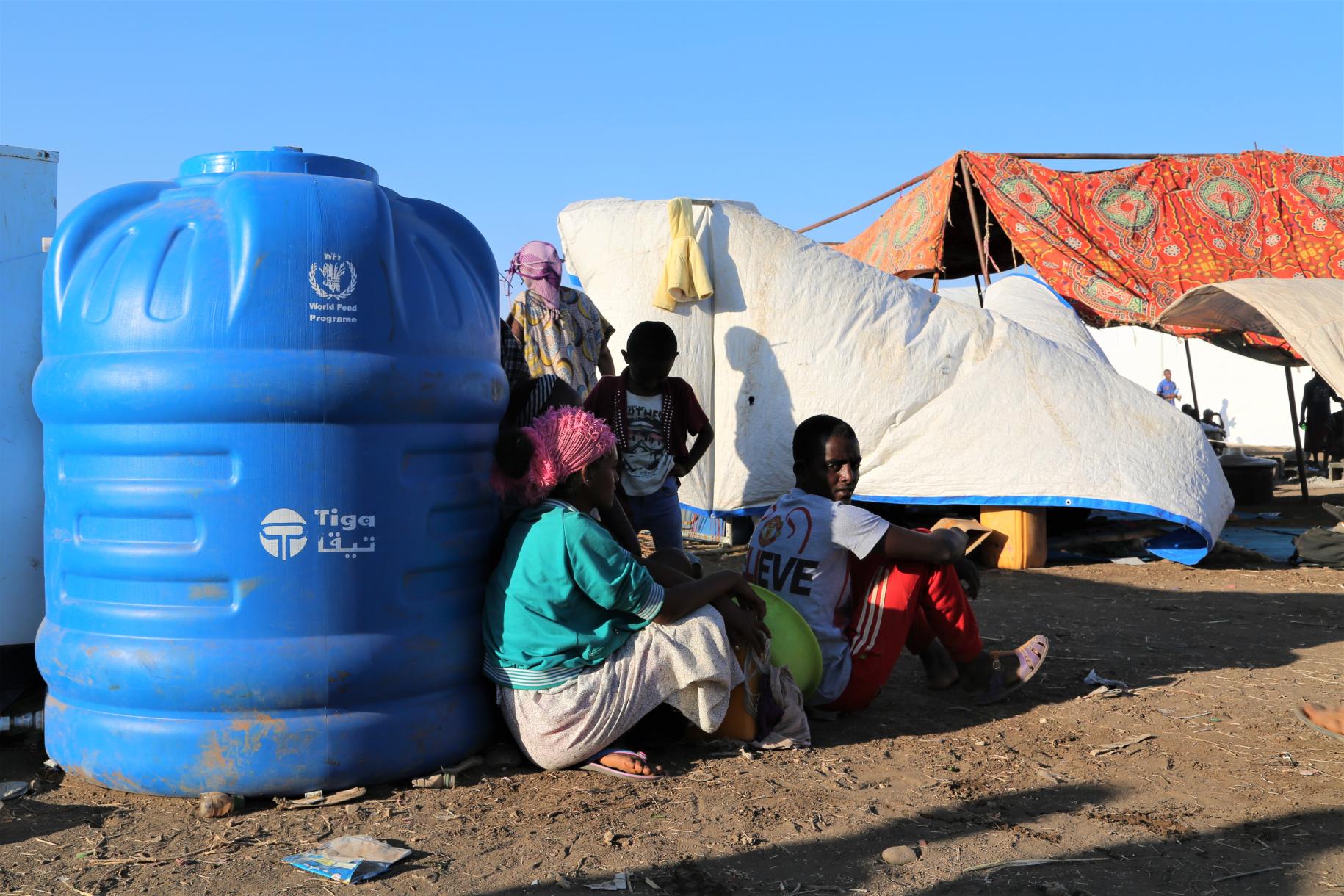 People sit against a large blue container of water at the camp.