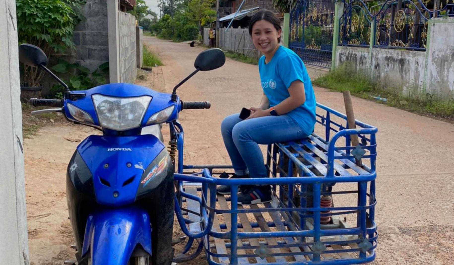 Une volontaire des Nations Unies vêtue d'un jean bleu et d'un t-shirt bleu portant le logo de l'UNICEF est assise dans le "side-cart" d'une moto bleue stationnée dans un chemin de terre et sourit à la caméra.