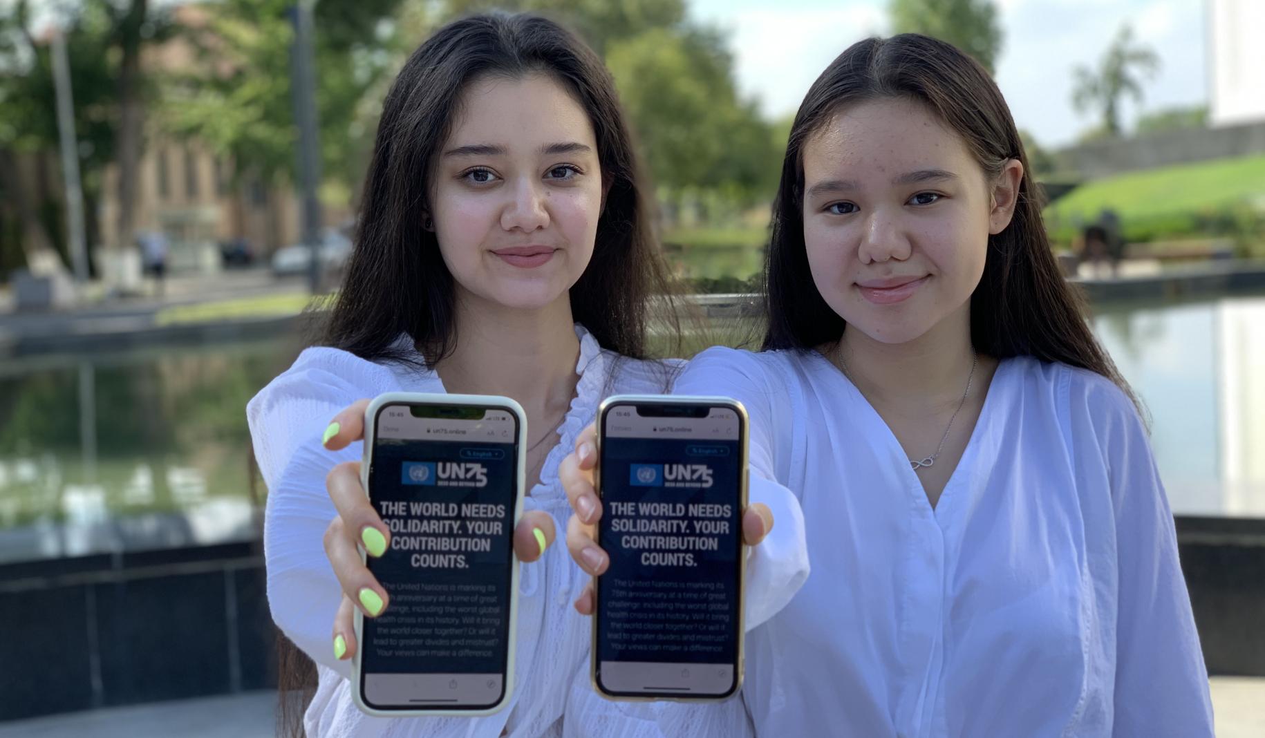 Two adolescent girls hold up their mobile phones directly towards the camera as they show the UN75 survey on the screen.