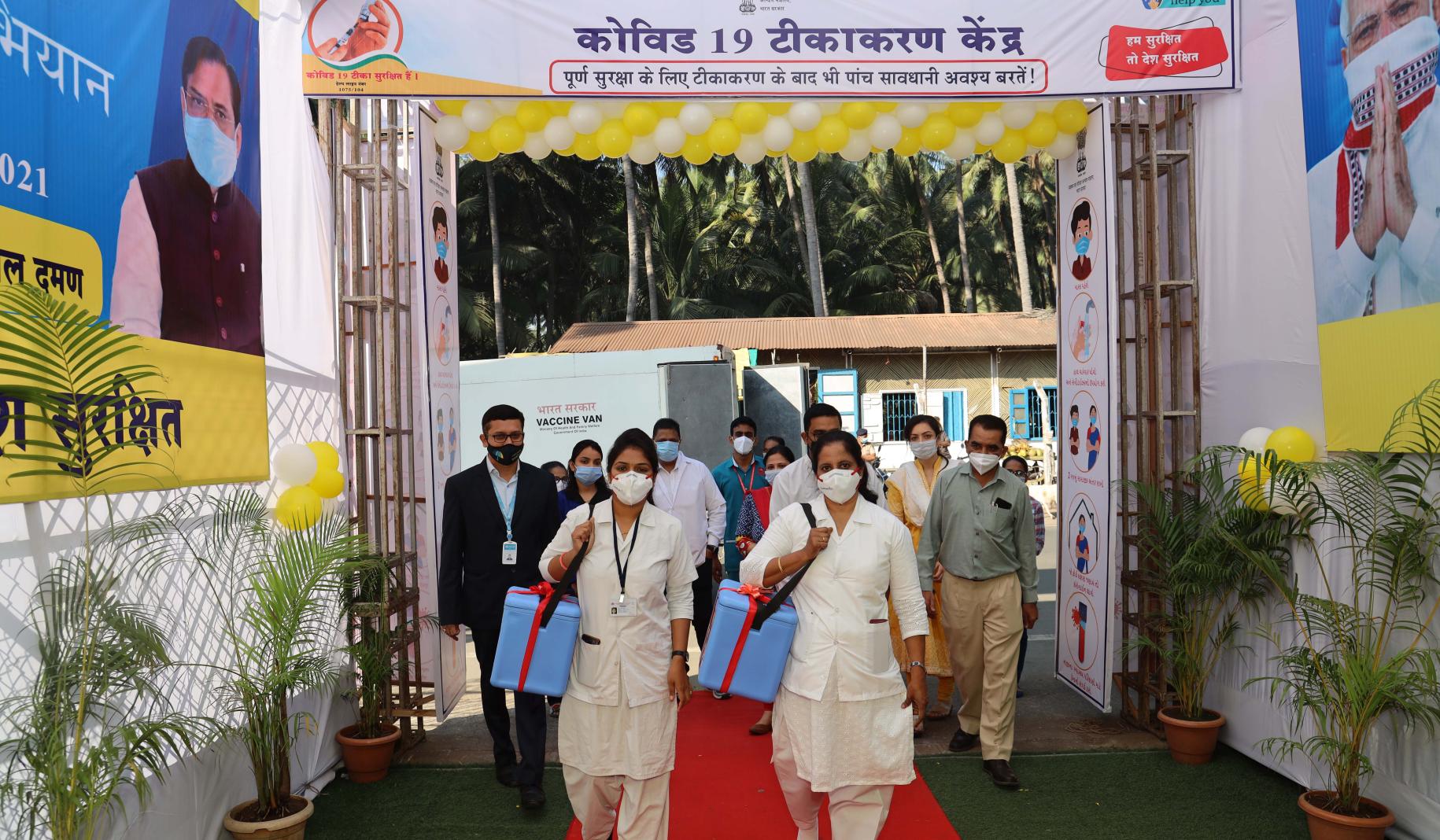 Two female healthcare workers wearing face masks walk towards the camera as they walk on a red carpet carrying vaccines as a group of people also wearing face masks follow behind them. 