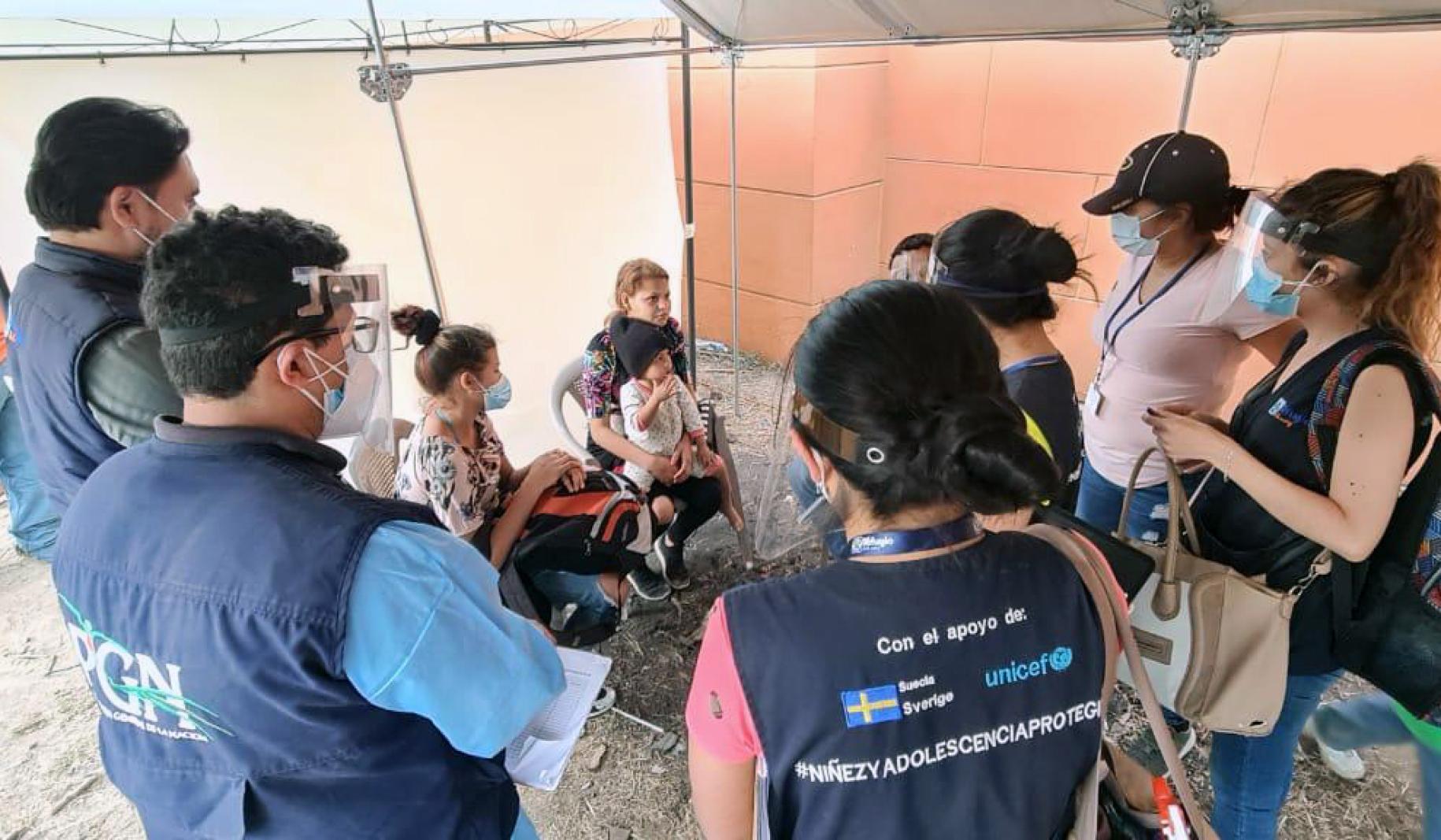 Personal de la ONU con mascarillas y protectores faciales rodean a una familia sentada en sillas en un círculo, todos se encuentran bajo una tienda de campaña.