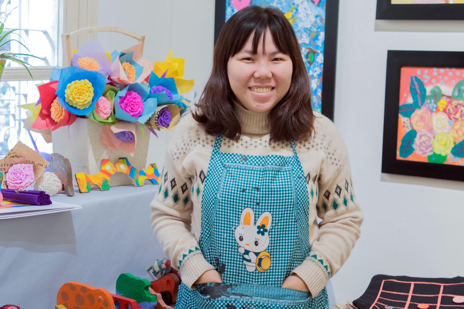 Tu Thanh Thuy smiles brightly at the camera as she stands with her hands in the pockets of her apron in front of colourful paper flowers and paintings.