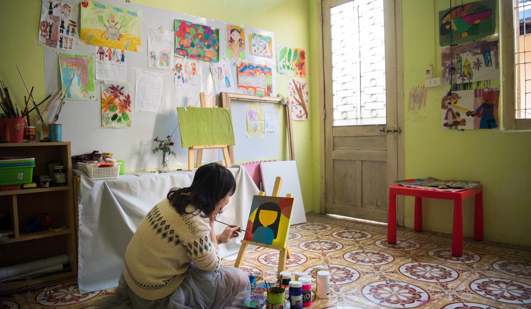 Tu Thanh Thuy sits on the floor of an art space and paints a picture placed on a small canvas.