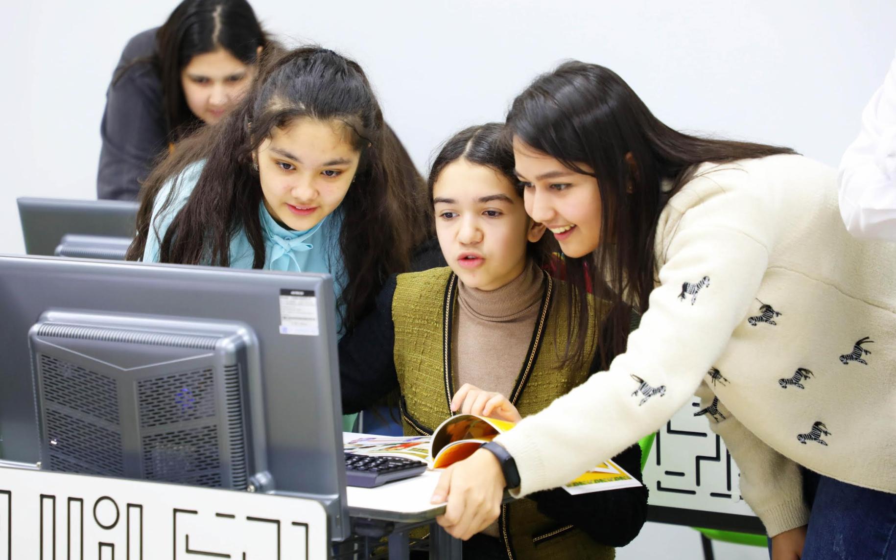 Группа улыбающихся девушек вместе работают за компьютером. Еще одна девушка на заднем плане работает за компьютером. 
