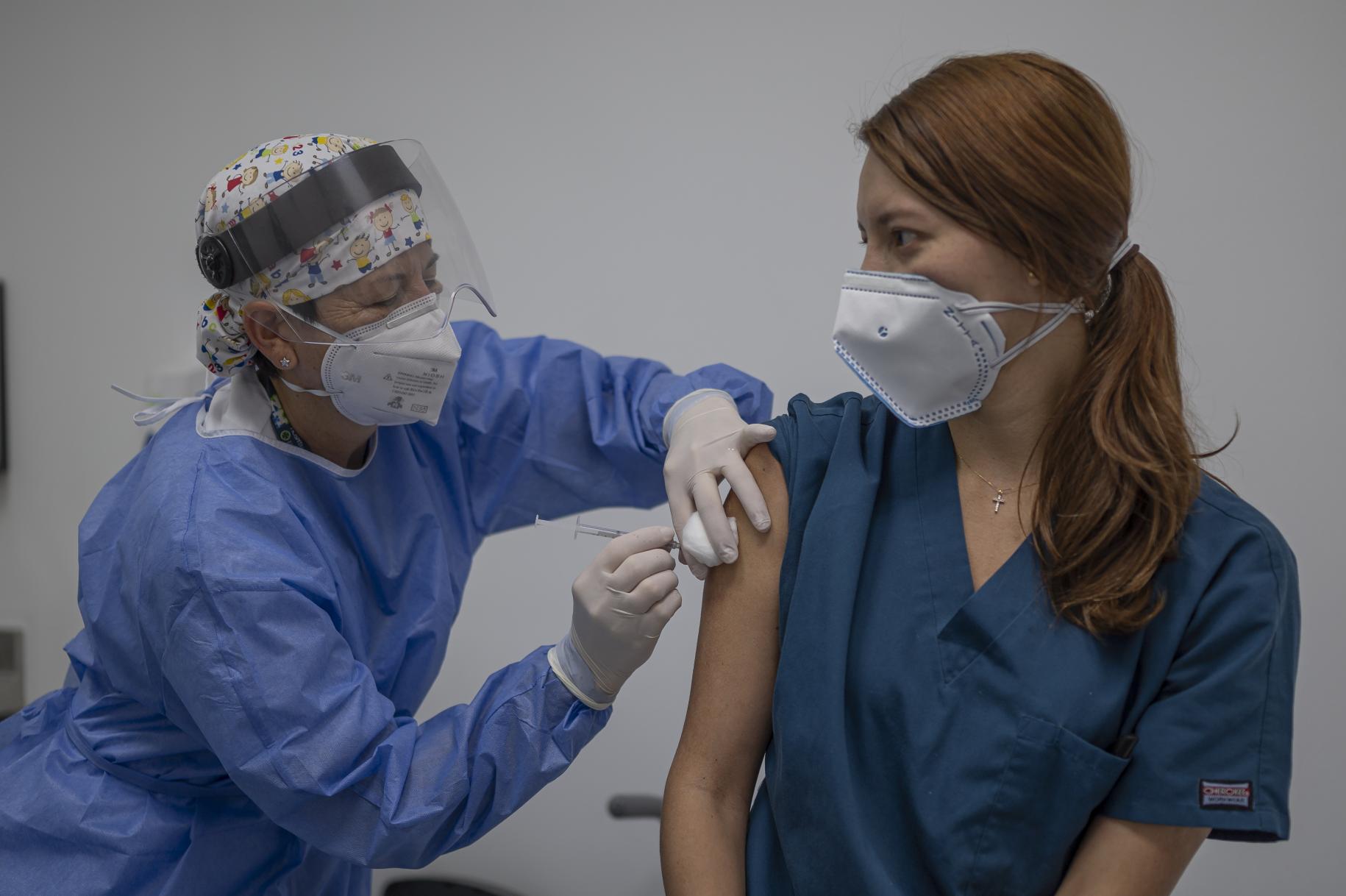 A healthcare professional wearing full head-to-toe protective gear is shown vaccinating a female colleague. 