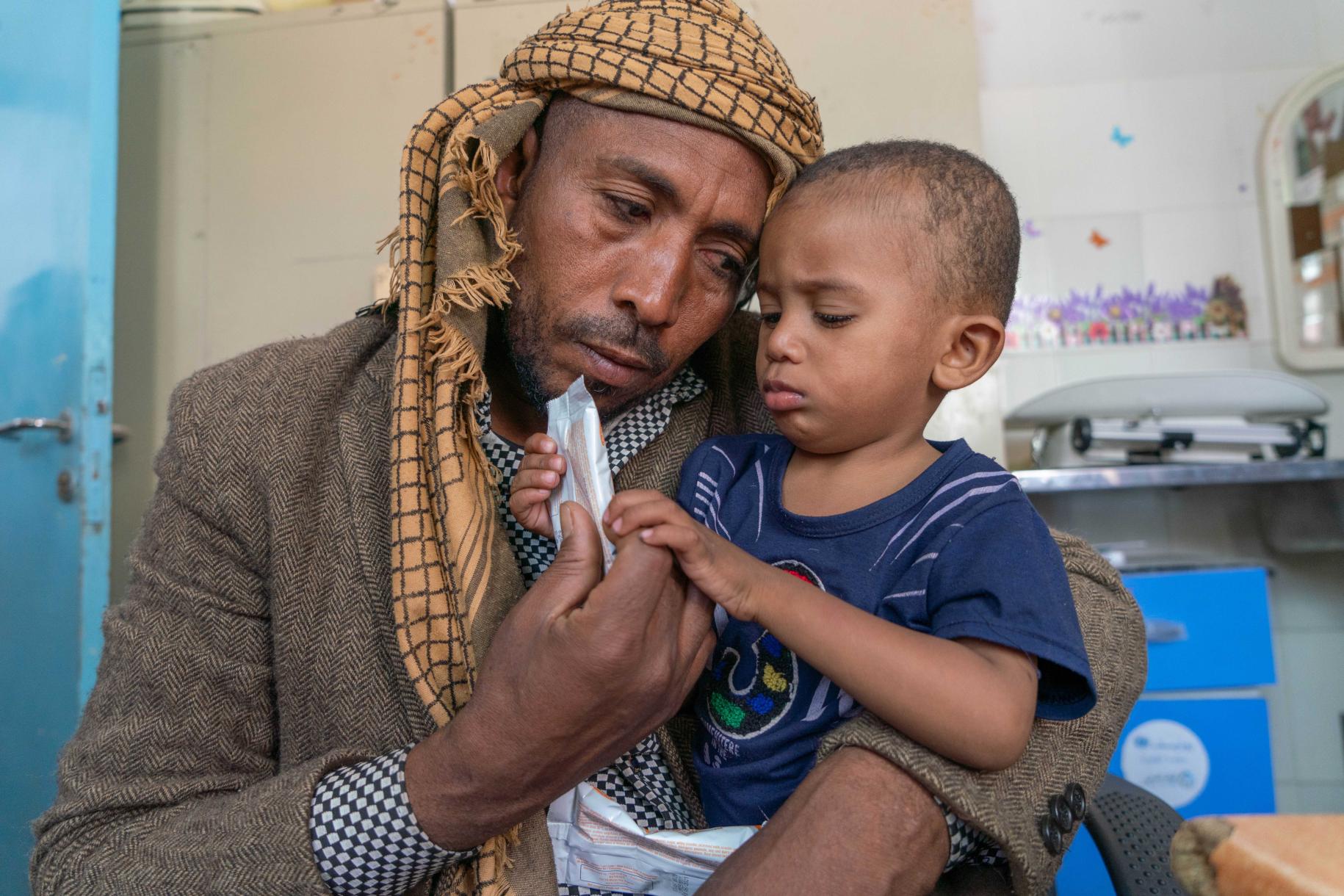 Sultan (1 año y 9 meses) que sufre de desnutrición moderada recibe suministros de un suplemento nutricional (Plumpy sup) en la clínica de salud apoyada por el PMA, en Amant Al Asimah. El niño pequeño aparece en primer plano, entre los brazos de su padre, Arafat (37), quien mira con amor a su hijo.
