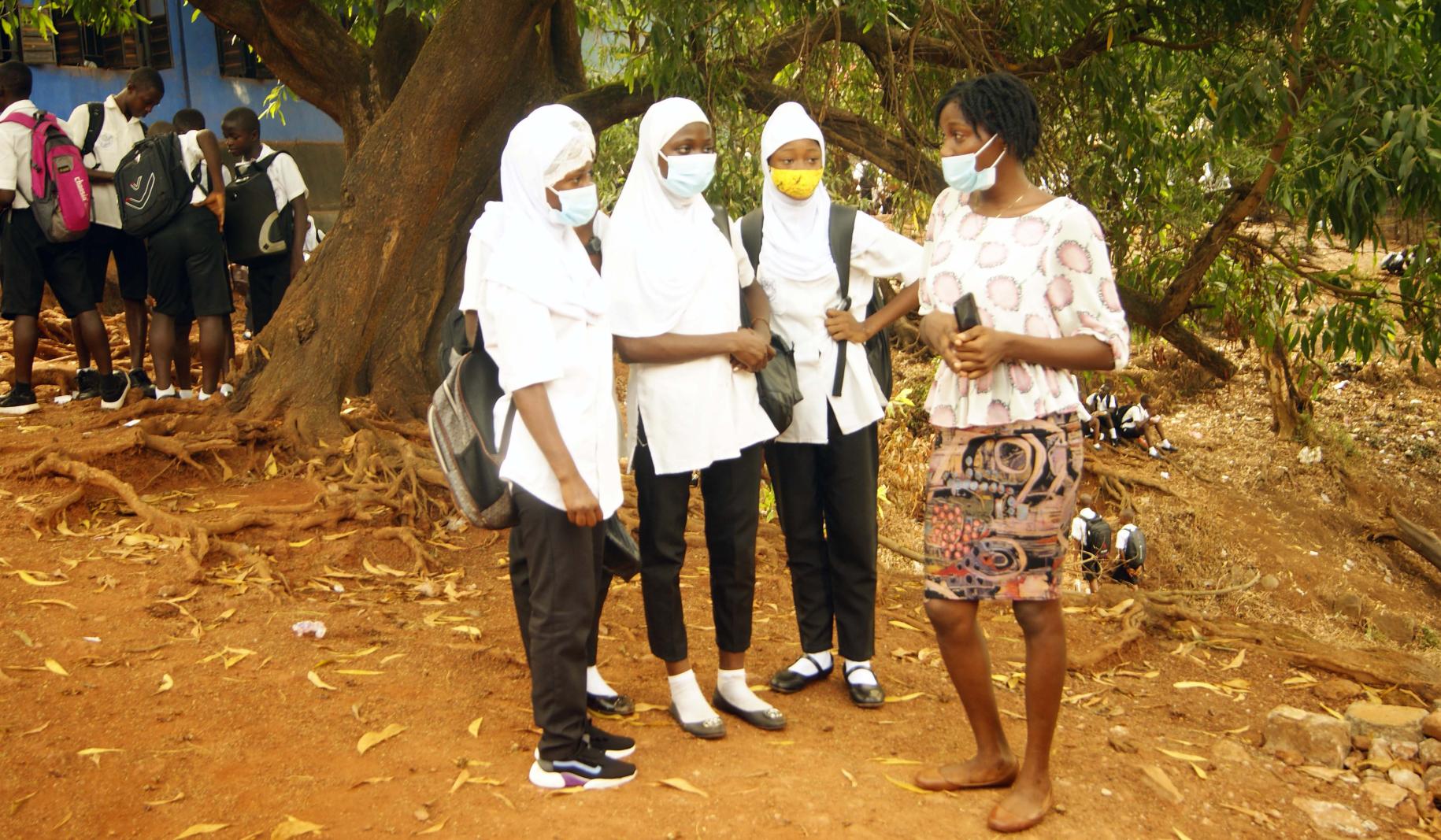 A woman wearing a face mask stands and speaks with three adolescent girls.