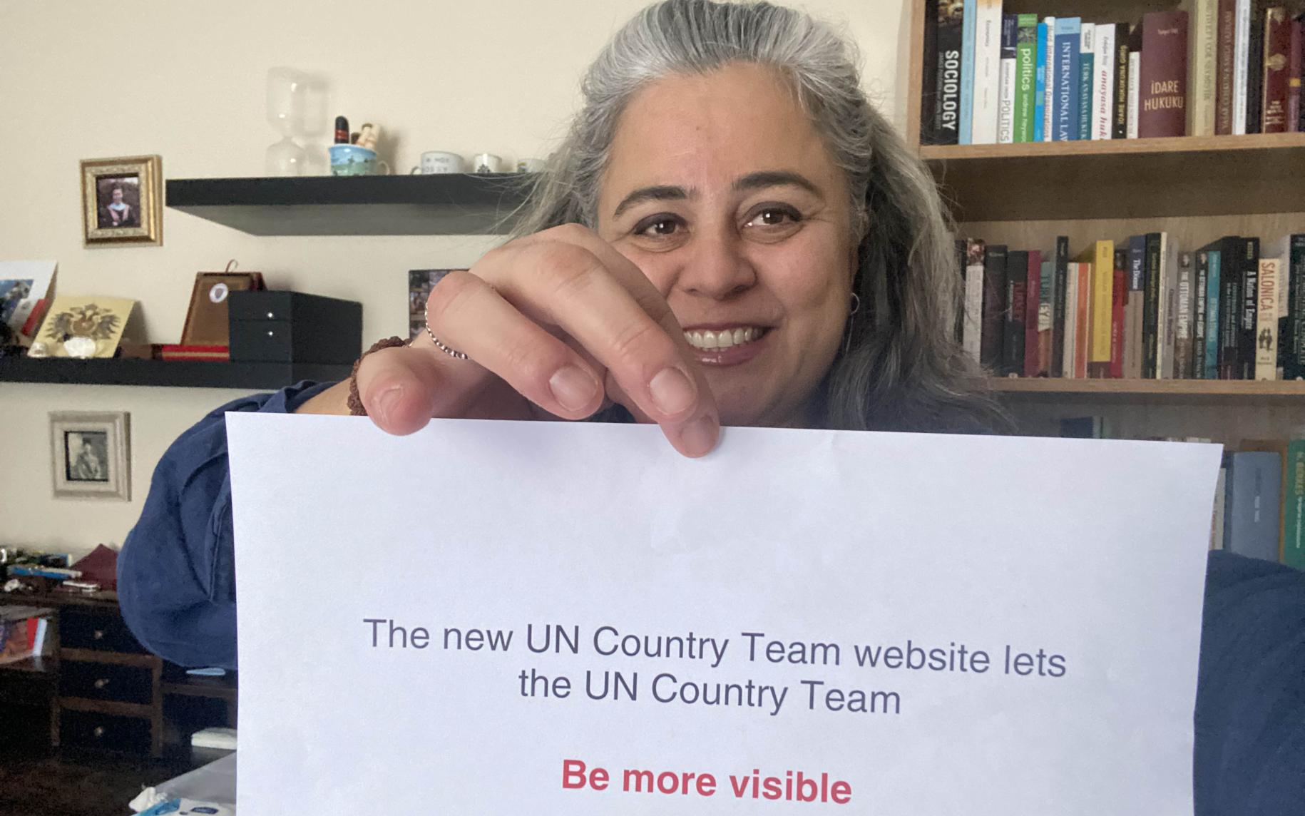 Una mujer sonríe alegremente a la cámara mientras sostiene un letrero que dice: "Los nuevos sitios web del equipo de país de la ONU permiten que el equipo de país de la ONU sea MÁS VISIBLE".
