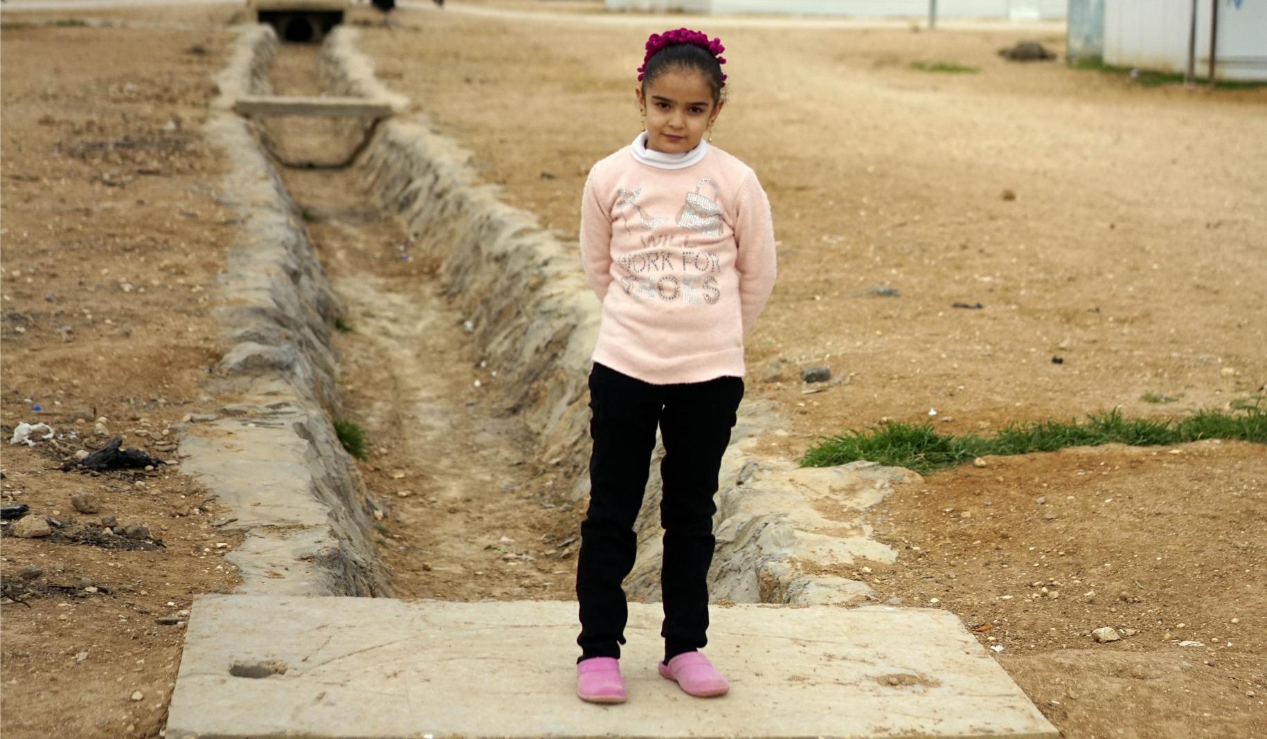 Laila, 10 años, con una tiara rosa y un sweater y zapatos en combinación, está parada afuera, frente a su casa. Parada, con sus manos detrás de la espalda, mira con determinación a la cámara.