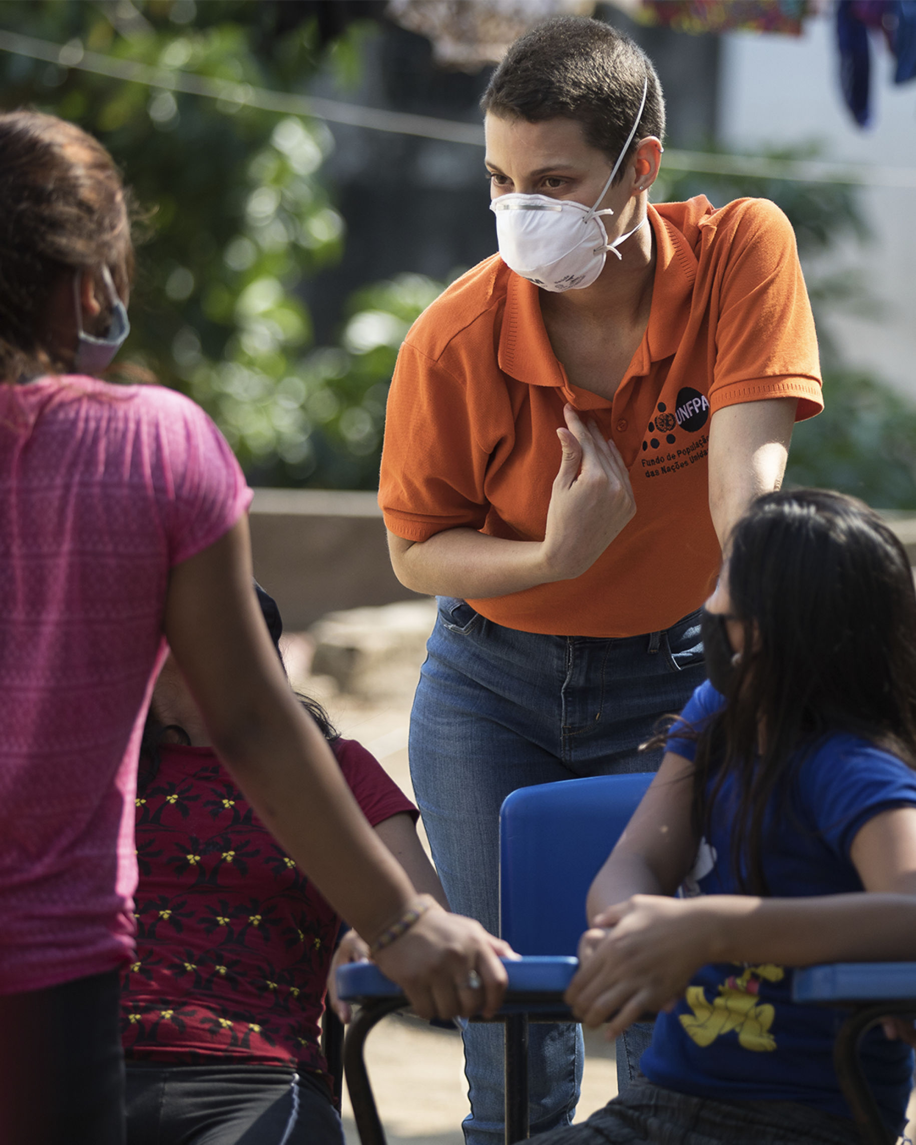 A woman in an orange shirt and a white face masks speaks to a group of women and girls. 