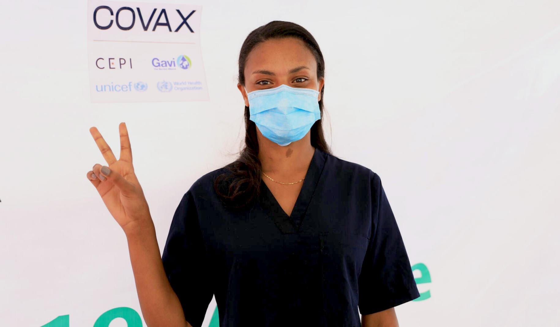 A healthcare professional stands by a COVAX sign and flashes a 'v' with her fingers to signify she was vaccinated.