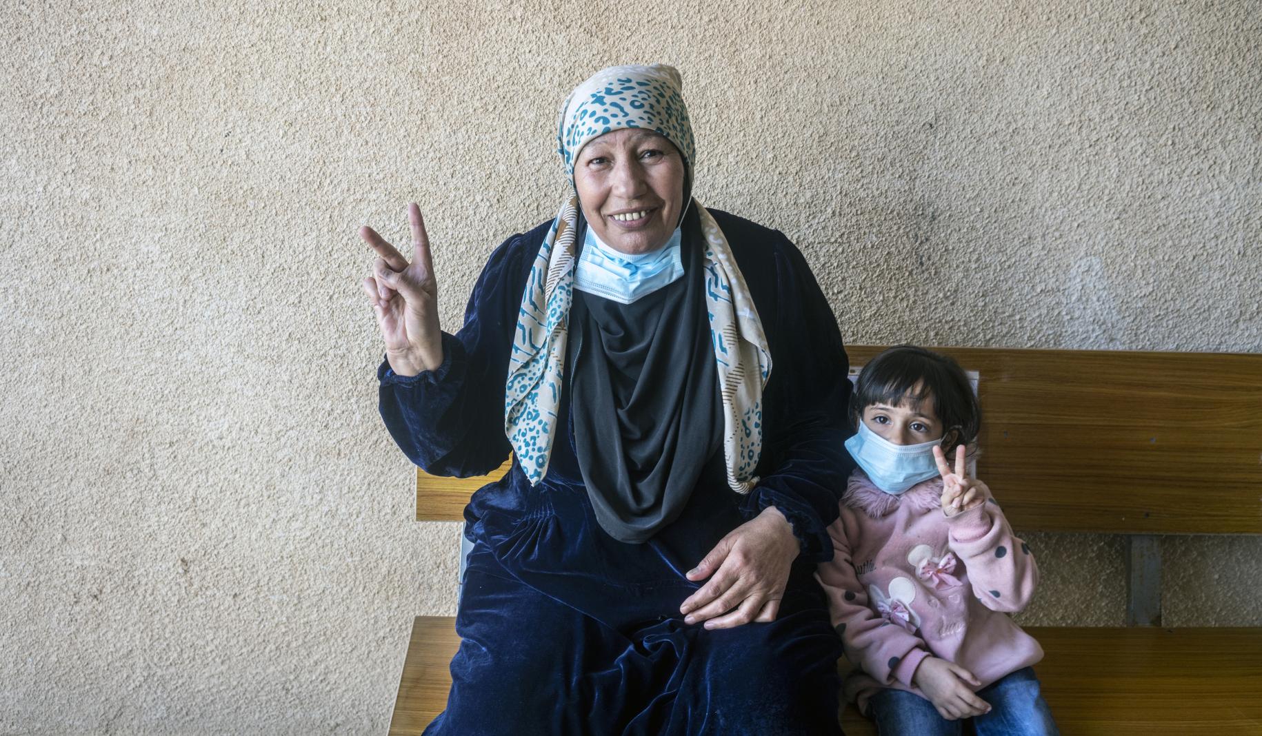 امرأة تبتسم للكاميرا مع حفيدتها الصغيرة. كلاهما يرفع علامة النصر بعد تلقي لقاح كوفيد-19.
